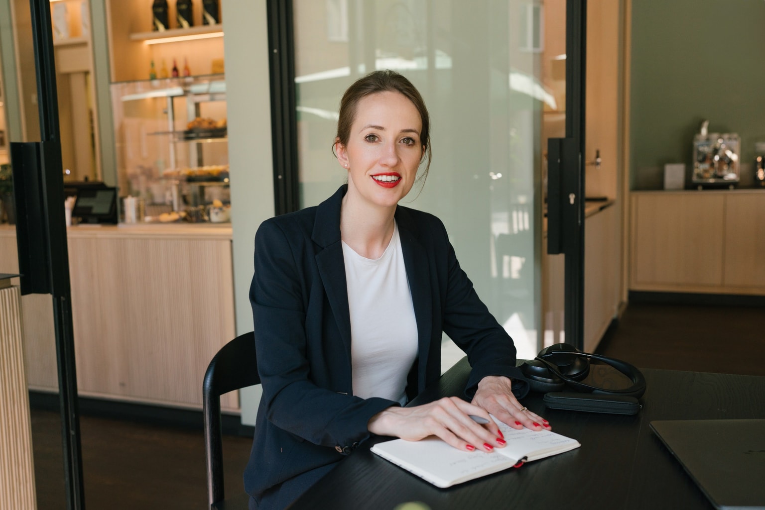 Woman in a blazer sits at a table with an open notebook and headphones in a modern cafe setting.