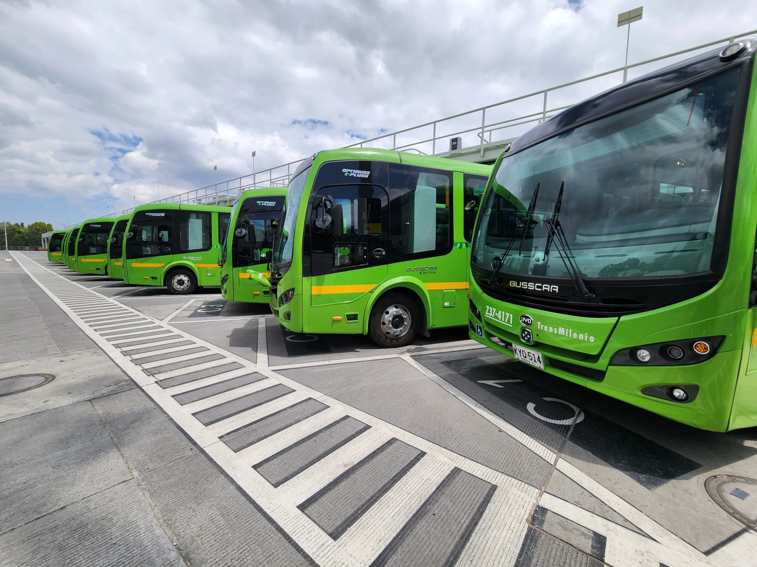 Plusieurs bus urbains verts garés en file sur une aire en béton sous un ciel nuageux, avec la marque et la signalétique des bus visibles.