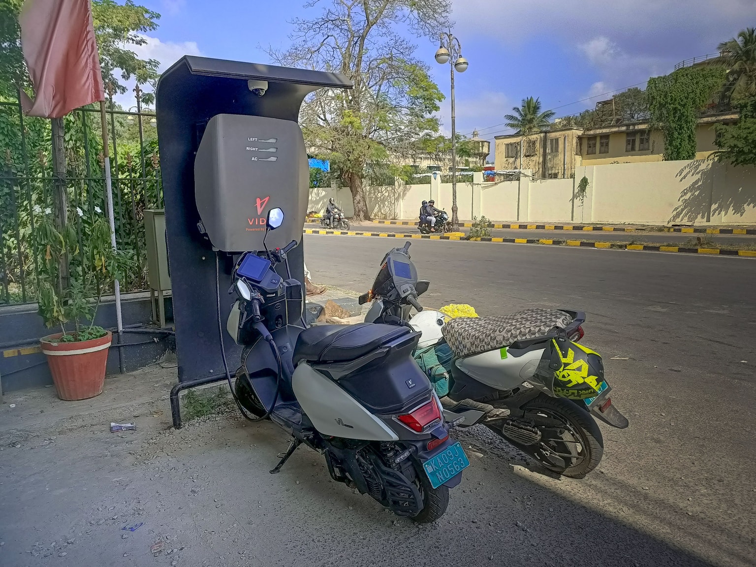 Two electric scooters parked and charging at a roadside charging station on a sunny day, with trees and buildings in the background.