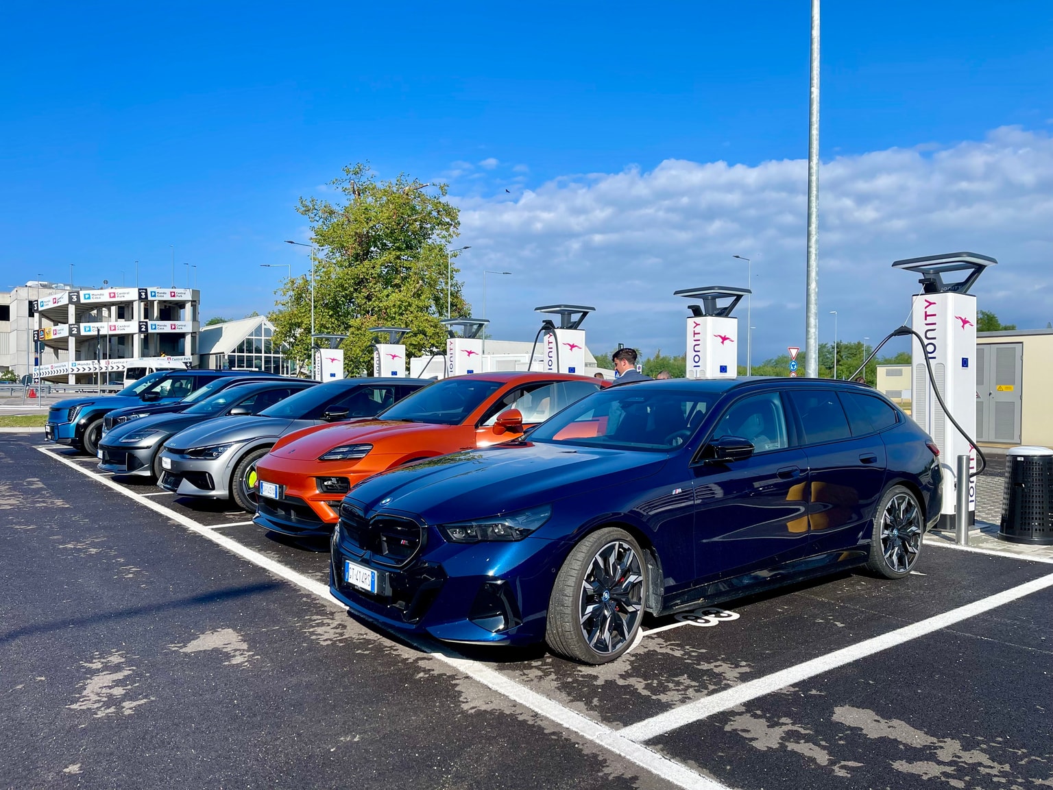 Una fila di auto elettriche colorate in ricarica presso una stazione di ricarica IONITY sotto un cielo azzurro, con un edificio e alberi sullo sfondo.