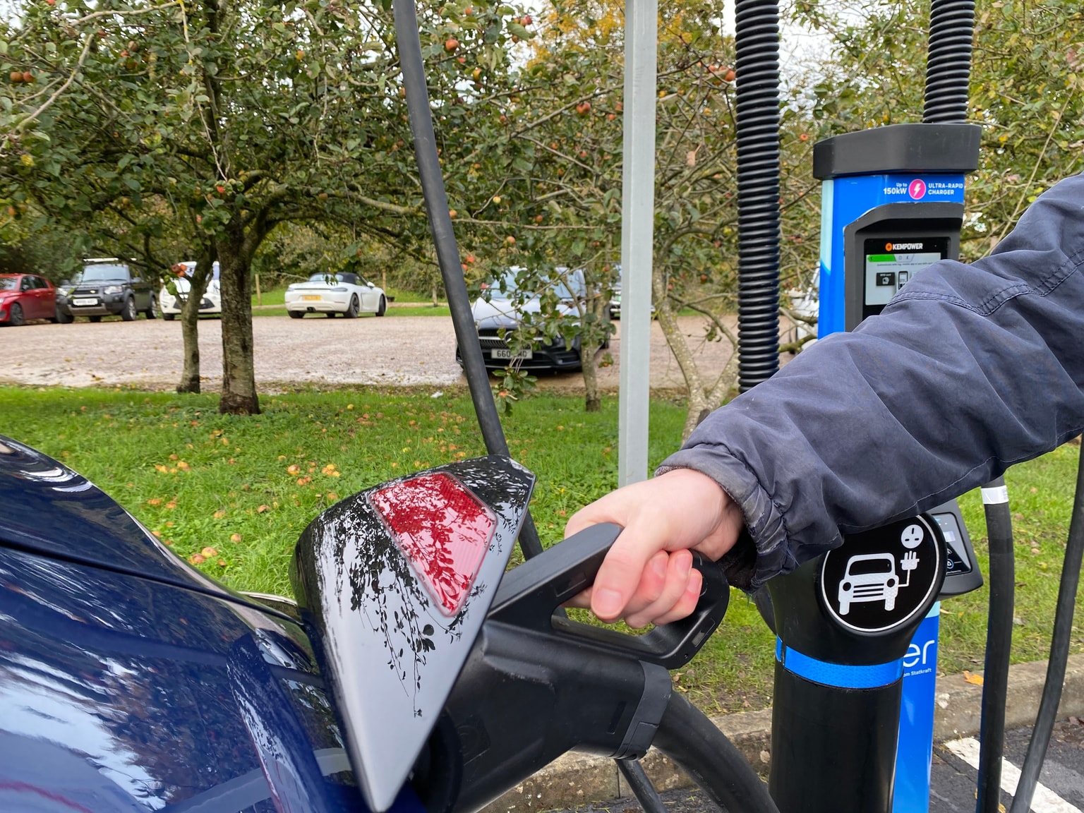 Una mano che tiene un cavo di ricarica collegato a un caricabatterie Mer con vegetazione sullo sfondo.