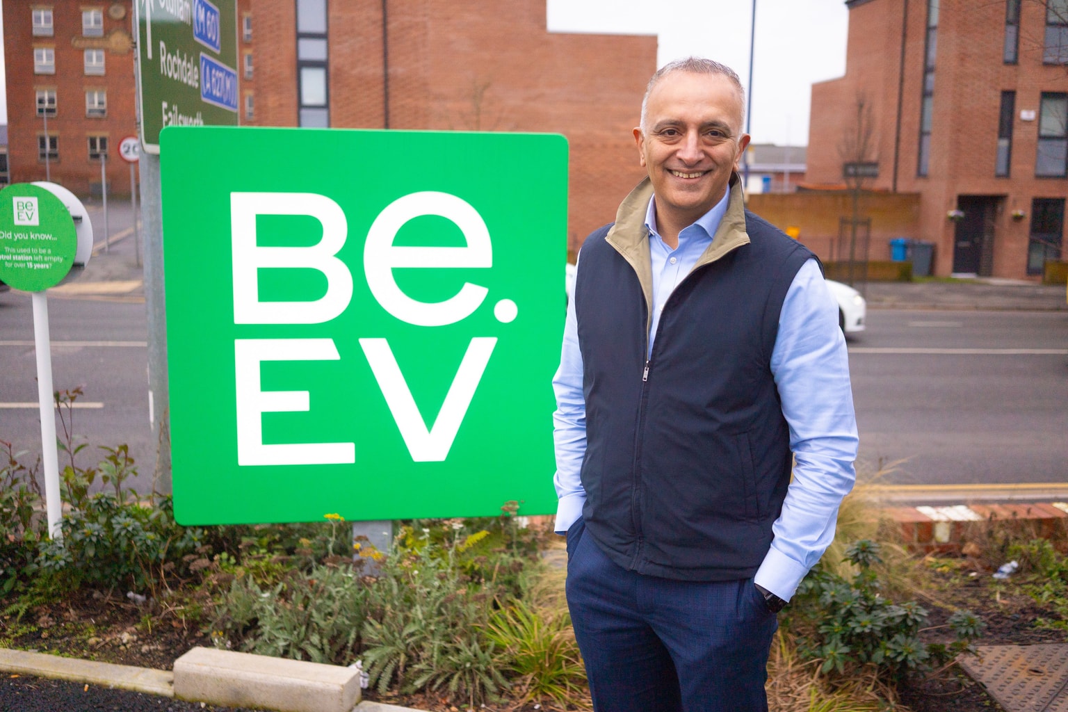 Man smiling next to a green "BE.EV" sign on a city street, with brick buildings in the background.