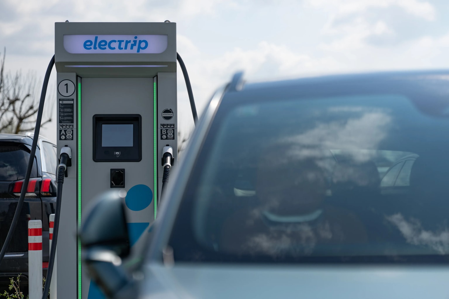 Stazione di ricarica per veicoli elettrici con il logo "electrip", parzialmente nascosto da un'auto, su sfondo di cielo nuvoloso.