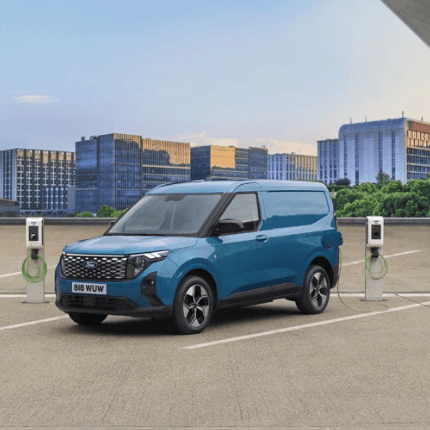 Blue electric van charging at a station in a city parking lot, with modern buildings and a clear sky in the background.