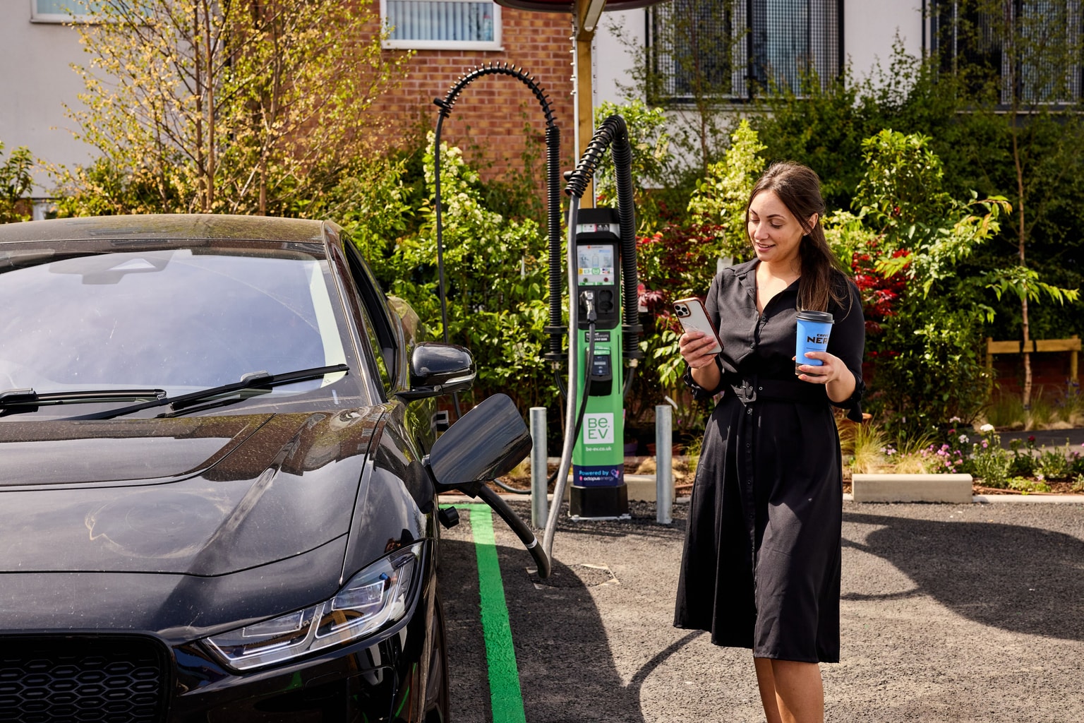 Woman in a black dress stands by an electric car at a Be.EV charging station, holding a phone and a coffee cup.