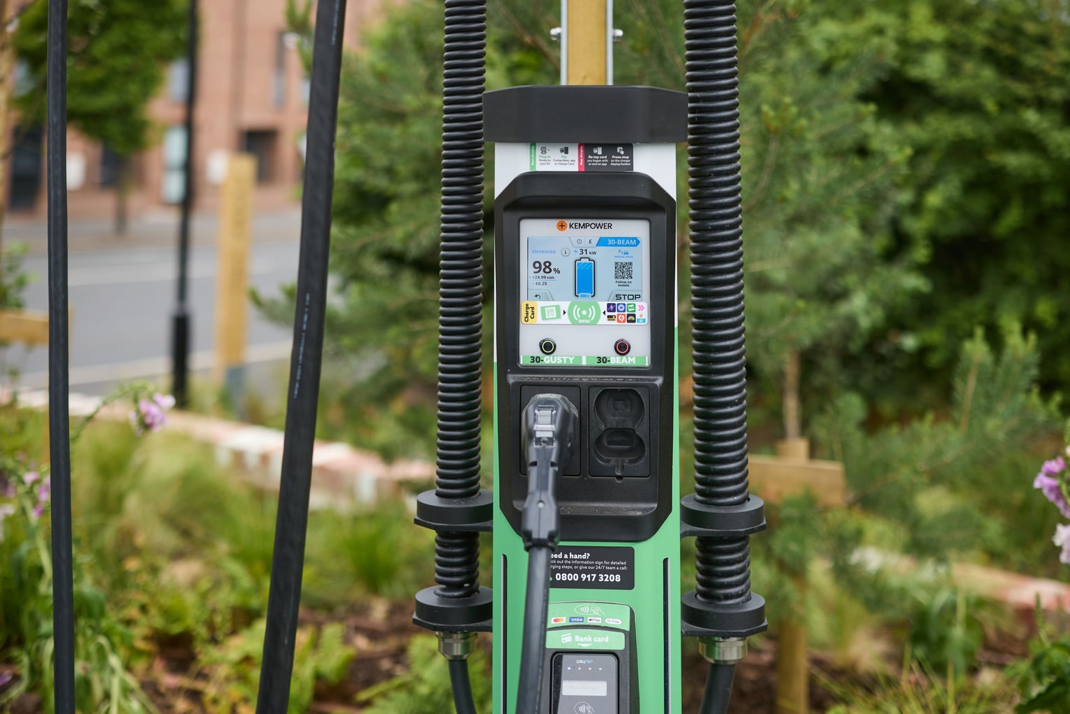 Close-up of a green Be.EV electric vehicle charging station with a screen displaying a charging progress of 98%, set outdoors with greenery.