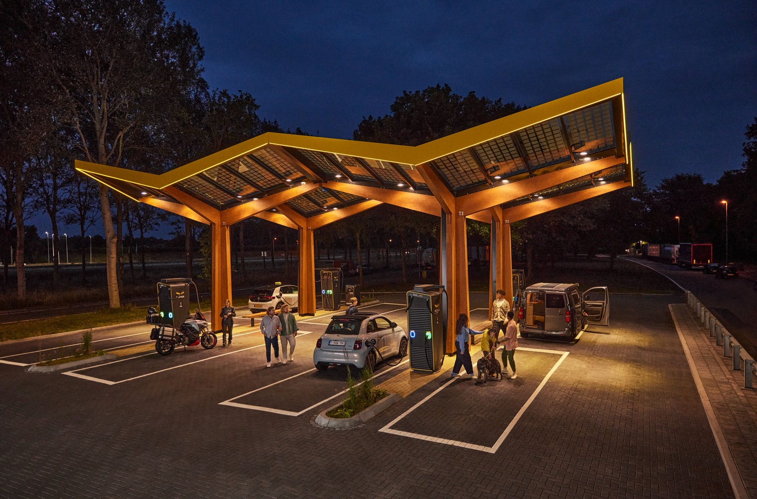 Station de recharge rapide pour véhicules électriques, ouverte de nuit, avec plusieurs voitures et des personnes sous un auvent moderne et lumineux.