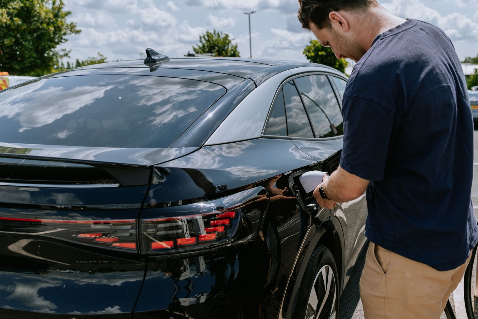 Man in a blue shirt charges a black electric car under a partly cloudy sky, with trees and cars in the background.