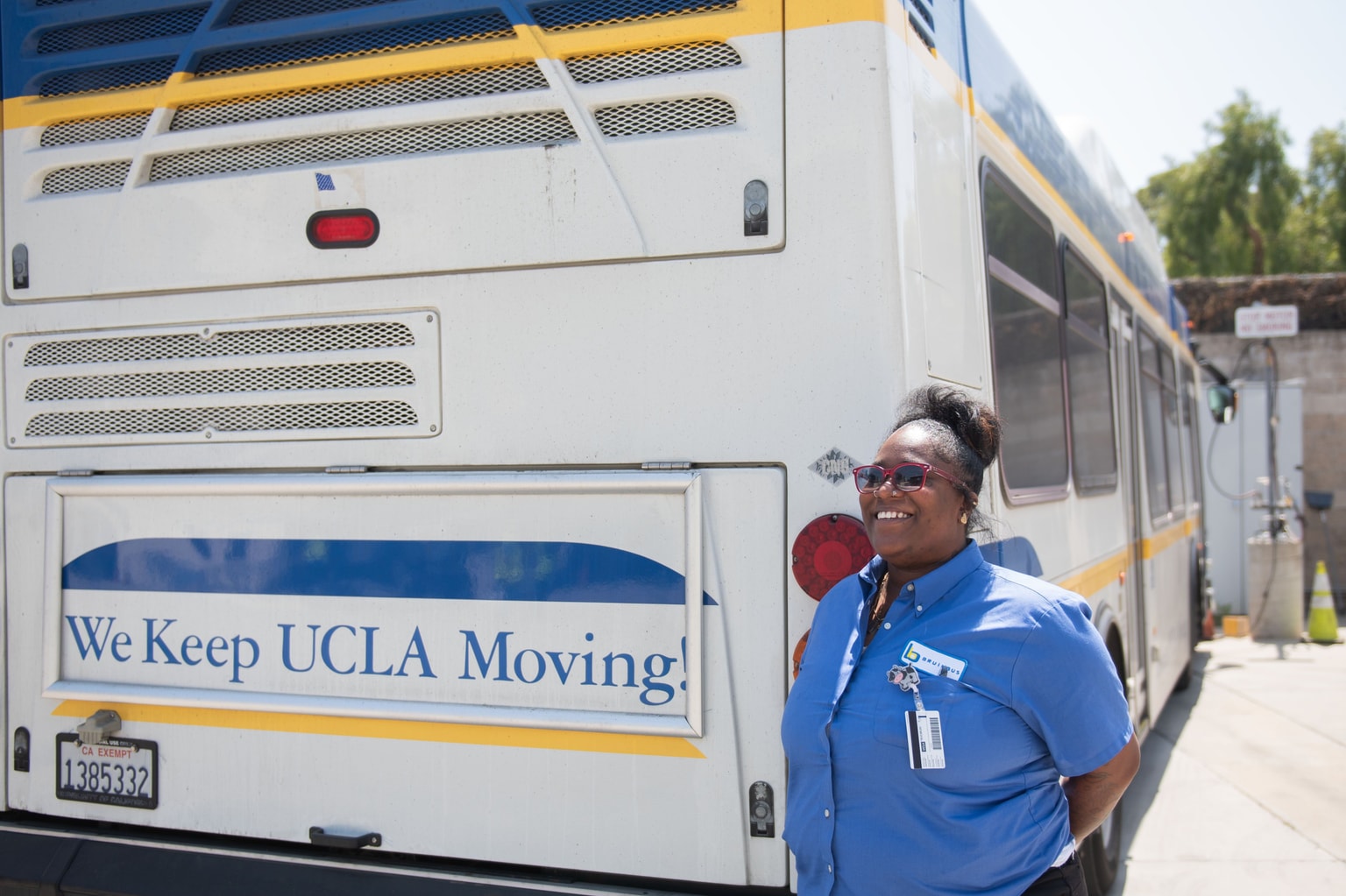 Un autista di autobus sorridente in uniforme blu è in piedi accanto a un autobus della UCLA con il cartello "We Keep UCLA Moving!"