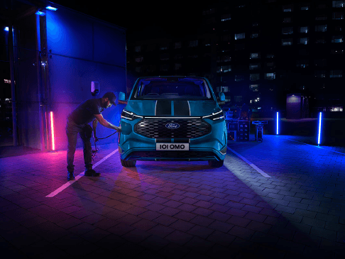 A person charges a blue Ford electric vehicle at a station, illuminated by vibrant blue and pink lights, with a cityscape backdrop at night.