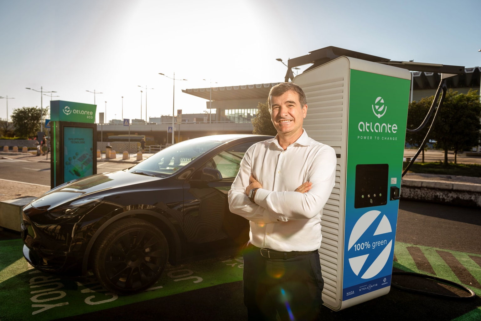Un uomo sorridente (Stefano Terranova) con una camicia bianca è in piedi accanto a un'auto elettrica nera presso una stazione di ricarica verde di Atlante energy, sotto un cielo soleggiato.