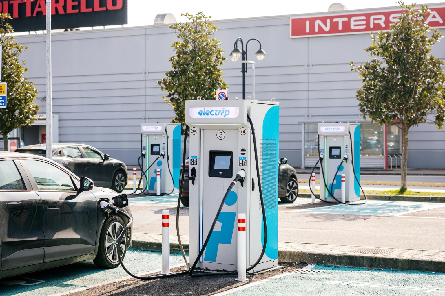 Un'auto elettrica è collegata a una colonnina di ricarica blu e bianca con la scritta "electrip", situata in un parcheggio dove si vedono altre colonnine sullo sfondo.