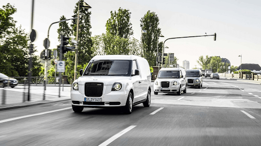 Taxis eléctricos blancos circulando por una calle urbana con árboles y semáforos al fondo.