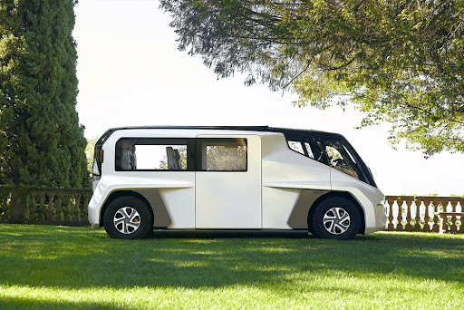 Vehículo autónomo blanco futurista aparcado sobre la hierba, rodeado de árboles y con una balaustrada de piedra al fondo.
