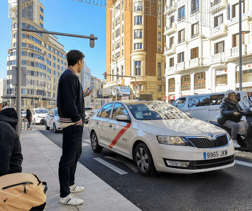 Un hombre de pie junto a una concurrida calle española, esperando un taxi blanco con una franja roja que lo atraviesa.