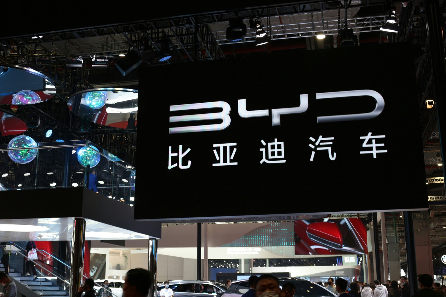 Large BYD sign with Chinese characters at an automotive show, surrounded by a busy crowd and reflective decorations.