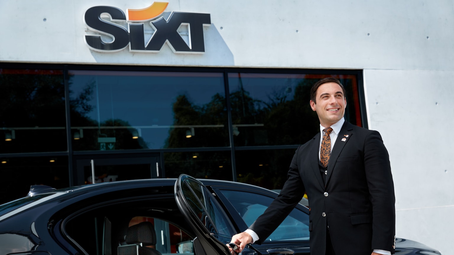 Uomo in giacca e cravatta che tiene la maniglia della portiera di un'auto, in piedi davanti a una sede di noleggio Sixt con un grande logo Sixt sopra.