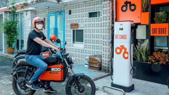 Persona su una motocicletta elettrica presso una stazione di ricarica, con casco e mascherina. La stazione reca il marchio “Dat Bike”.