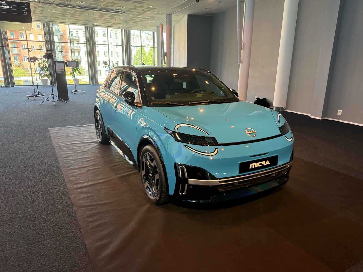 Una Nissan Micra blu è esposta all'interno, su una piattaforma nera, con grandi finestre e la città sullo sfondo.