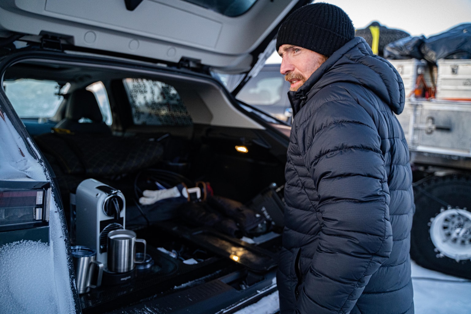 Un uomo con giacca nera e berretto è in piedi vicino al bagagliaio aperto di un'auto con una macchina per il caffè in una giornata nevosa.