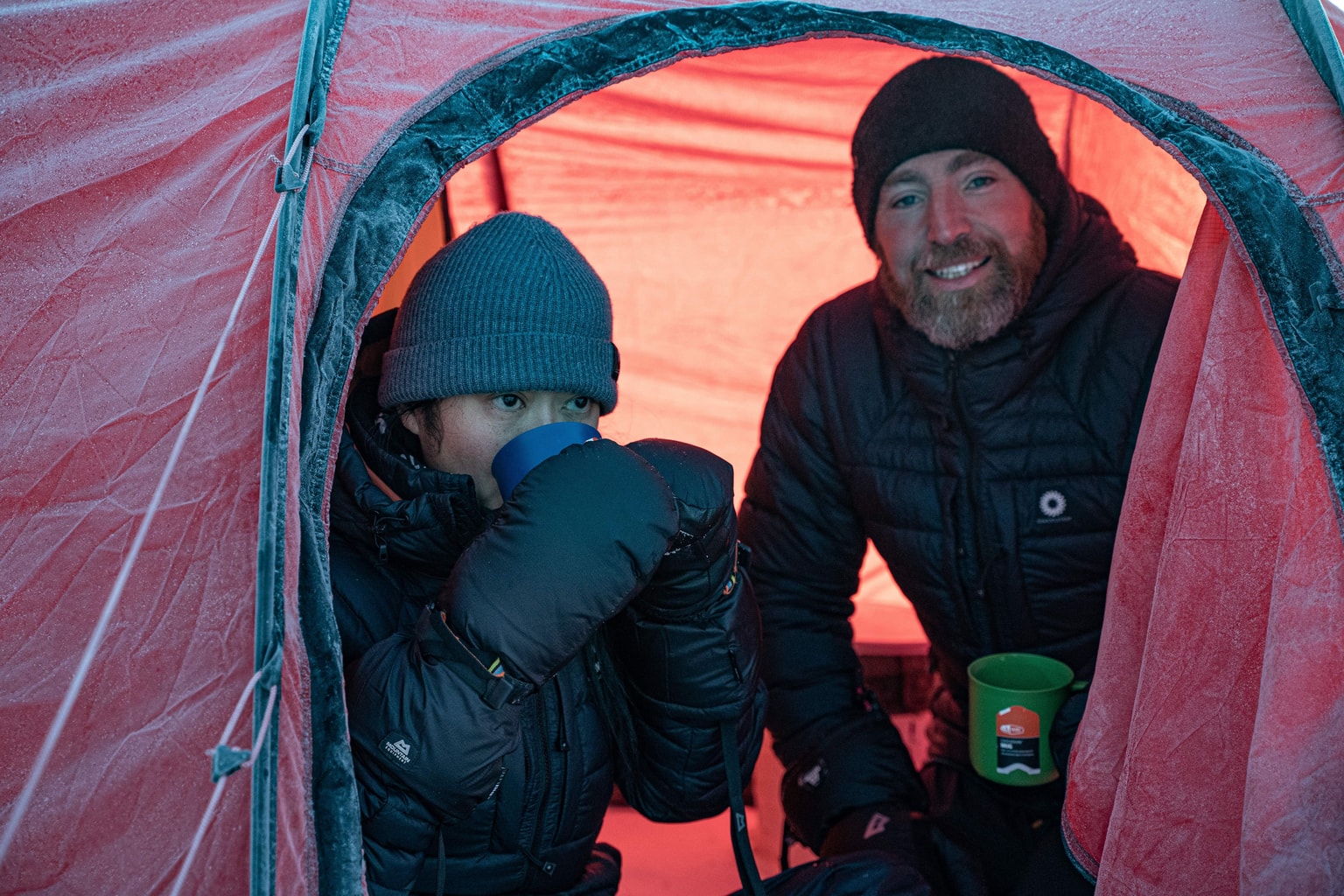 Due persone in abbigliamento invernale all'interno di una tenda rossa; una beve da una tazza, l'altra sorride, entrambe indossano cappello e guanti.