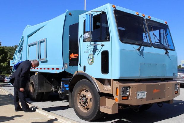 A large blue electric garbage truck parked on the street as a person in a suit examines something near the front wheel.