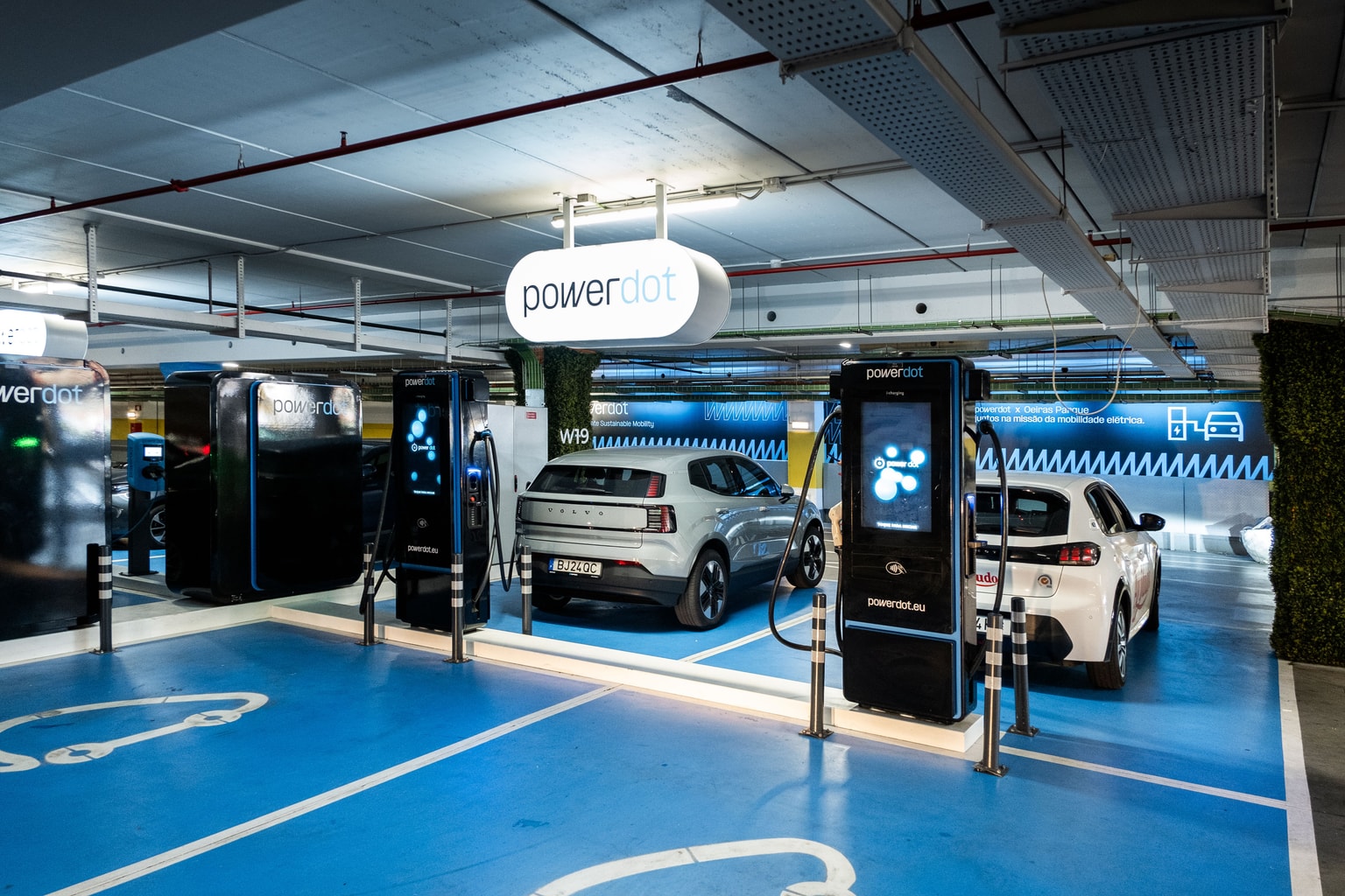 Veículos elétricos a carregar nas estações Powerdot num parque de estacionamento moderno e bem iluminado, com lugares marcados a azul para veículos elétricos.