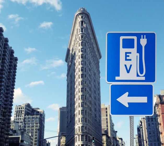Iconic flatiron building with an EV charging station sign pointing left in the foreground, clear blue sky.