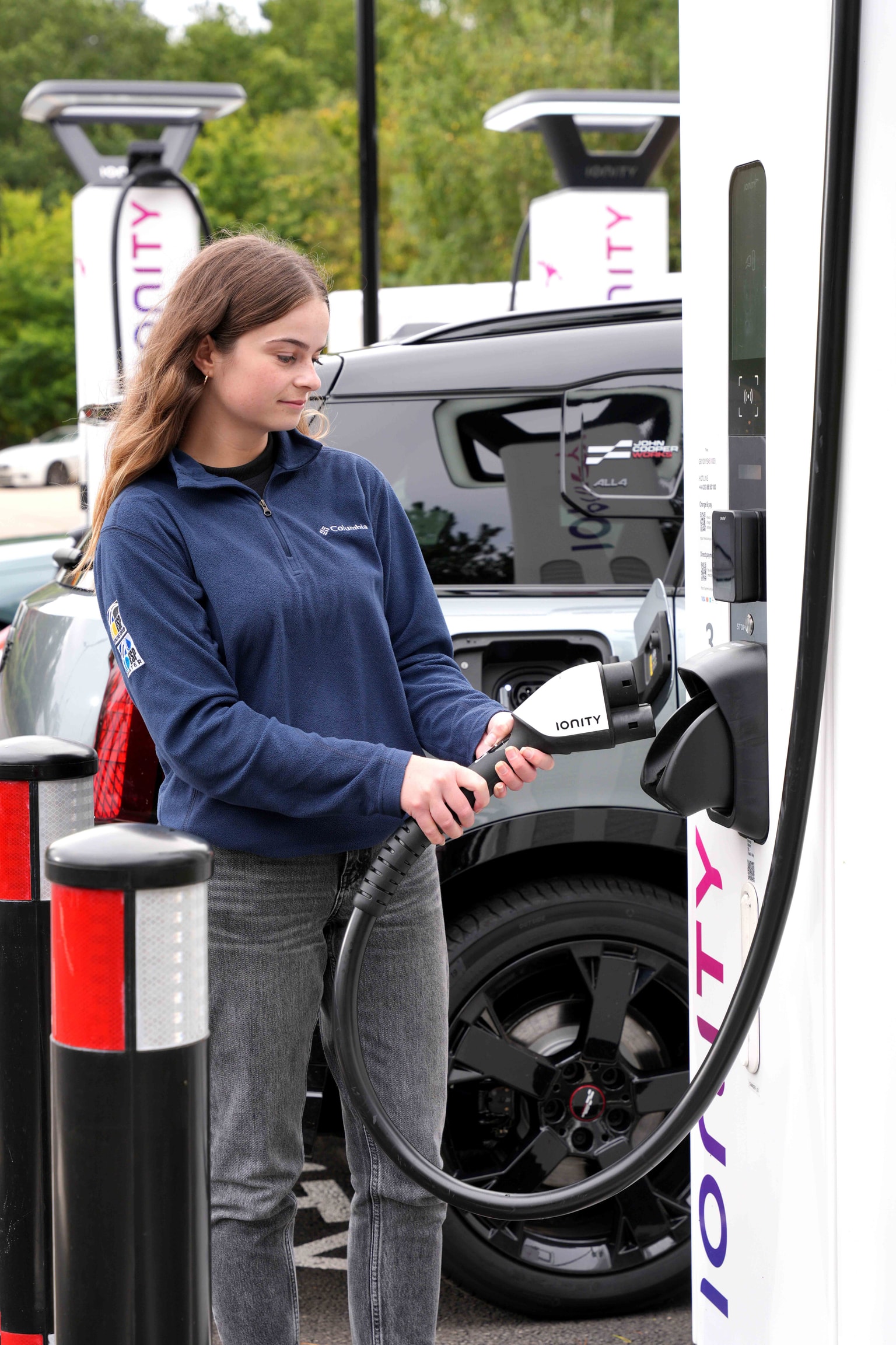 Una donna con una giacca blu ricarica un'auto elettrica in una stazione di ricarica IONITY. Tiene in mano la spina di ricarica, con auto e vegetazione sullo sfondo.