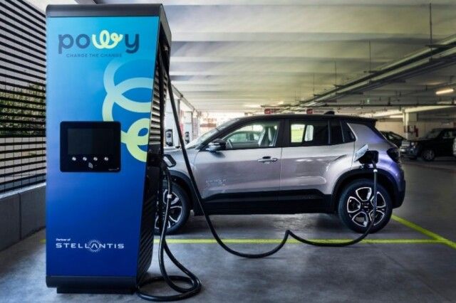 SUV elettrico in ricarica presso una stazione Powy blu in un parcheggio coperto, con cavi collegati, sotto luci fluorescenti.