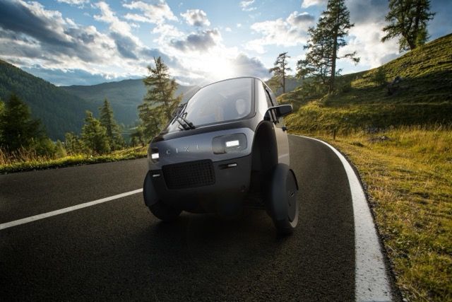 Futuristic three-wheeled vehicle driving on a scenic mountain road with trees and a cloudy sky in the background.