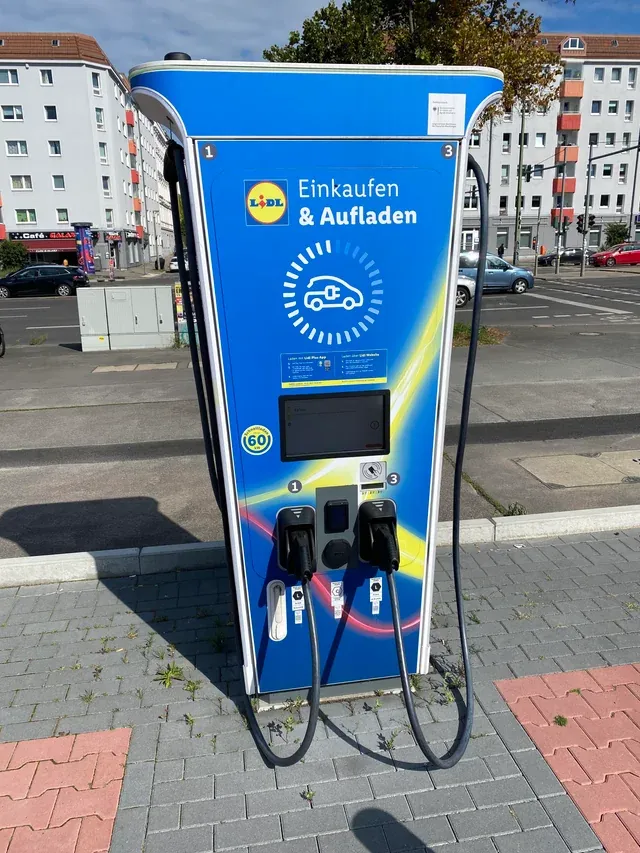 A blue electric vehicle charging station with "Lidl Einkaufen & Aufladen" written on it, located in an urban area with buildings in the background.