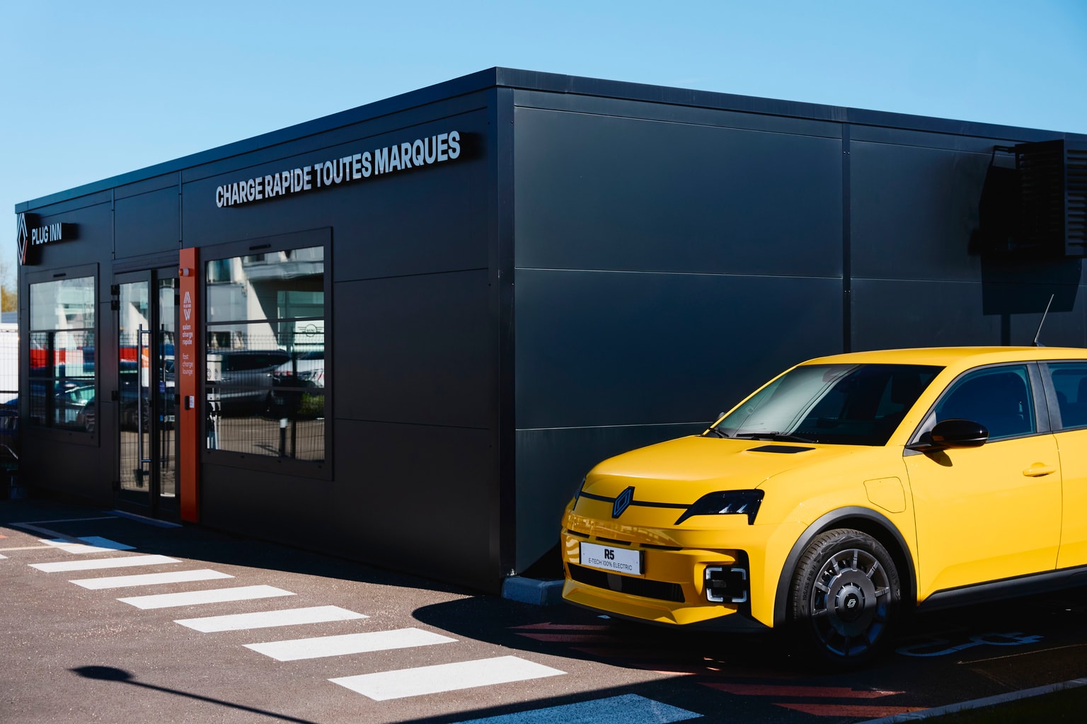A yellow electric car - the Renault 5 - parked outside a modern building with signage "Charge Rapide Toutes Marques" under a clear blue sky.