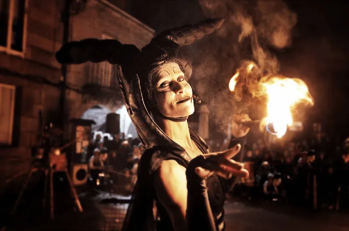 A performer in a jester costume with elaborate horns smiles while a flame burns in the background, surrounded by a nighttime crowd.