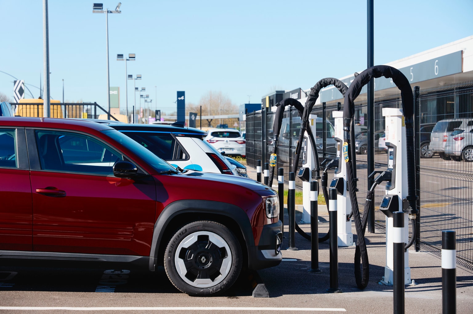 A red Renault is parked in front of Plug Inn Fast Charge chargers, with a blue sky background.
