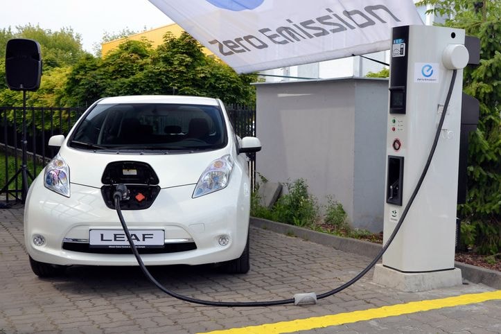 White electric Leaf charging at a station with a cable connected under a "Zero Emission" banner. Surrounded by greenery.