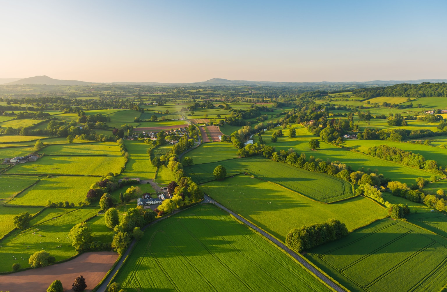Vue aérienne d'un patchwork de terres agricoles verdoyantes parsemées de haies, de fermes isolées et de collines ondulantes sous un ciel clair à l'heure dorée.