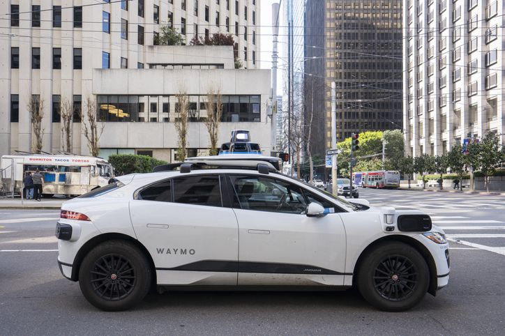 A Waymo self-driving car is parked on a city street with tall buildings in the background.