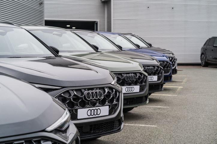 Reihe geparkter Audi-Fahrzeuge in verschiedenen Farben vor einem Gebäude mit Metallverkleidung, aufgenommen aus der Frontalperspektive.