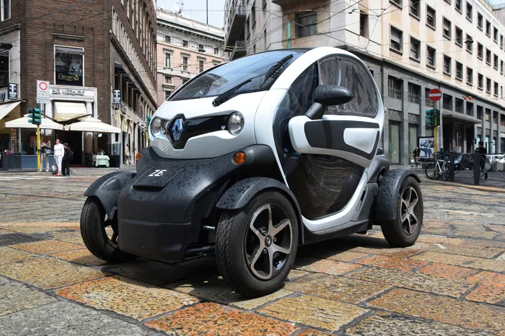 Ein kleines, futuristisches zweisitziges Elektrofahrzeug, geparkt auf einer Kopfsteinpflasterstraße in städtischer Umgebung, mit Gebäuden im Hintergrund.