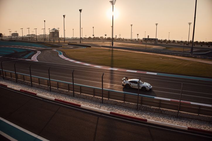 Race car speeding on a curving track at sunset, with floodlights and greenery in the background under a clear sky.