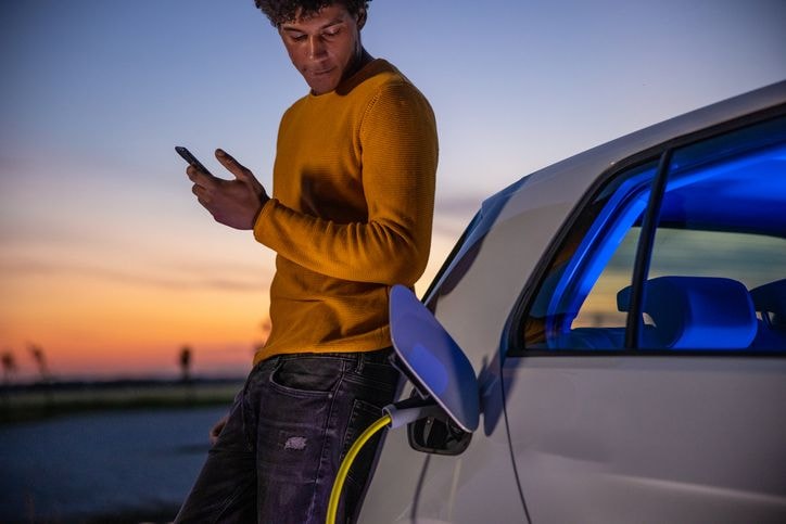 Man in yellow sweater using phone, leaning on electric car charging at dusk, with interior lit in blue.