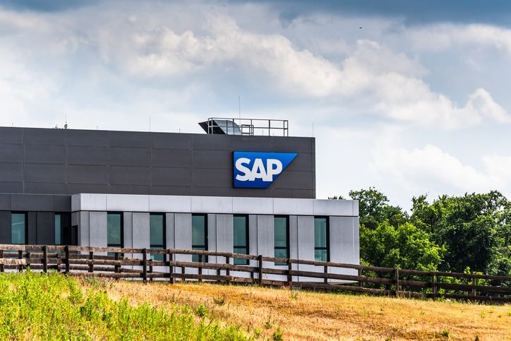 Modern building with "SAP" logo against a cloudy sky, surrounded by greenery and a wooden fence in the foreground.