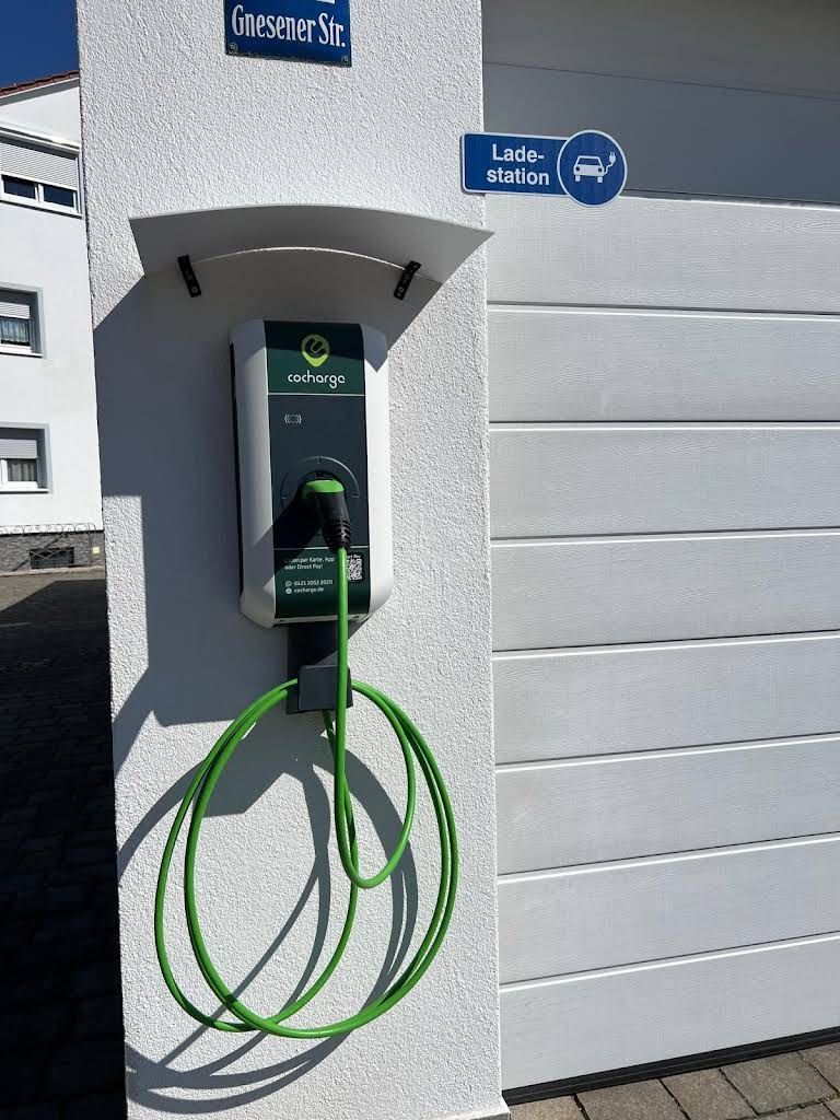 Cocharge-Ladestation für Elektrofahrzeuge mit einem grünen Kabel, montiert an einer weißen Wand, unter einem „Ladestation“-Schild.
