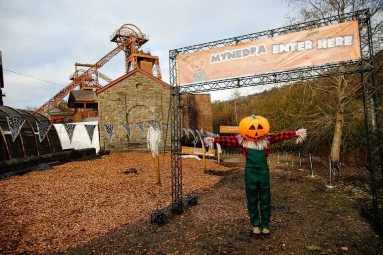 Pumpkin-headed scarecrow in overalls stands by an entrance sign at an outdoor event near an old industrial site with flags and festive banners.