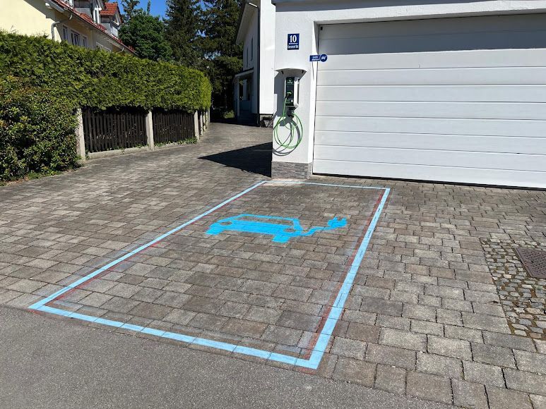 Auffahrt mit einer Cocharge-Ladestation für Elektrofahrzeuge, gekennzeichnet durch blaue Linien und ein Autosymbol, vor einer weißen Garage.