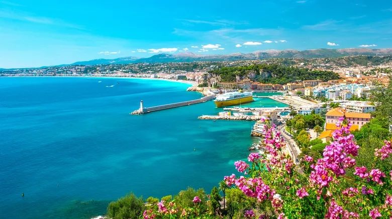 Paysage urbain côtier de Nice, France avec une mer bleue vibrante, un phare et des fleurs roses au premier plan sous un ciel bleu clair.