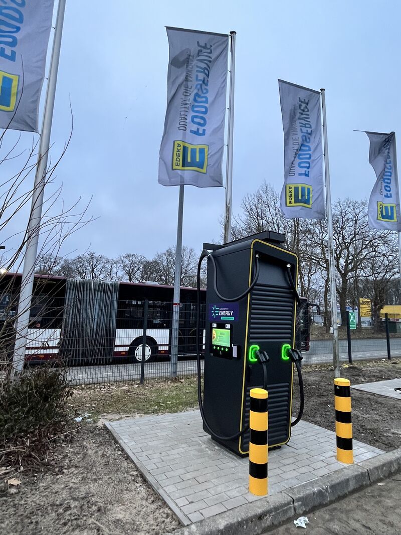 Electric vehicle charging station in a parking area, surrounded by yellow and black bollards, with flags and a bus in the background.
