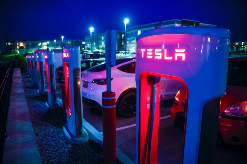 Stazione Tesla Supercharger di notte, con diverse auto parcheggiate e in ricarica. Luci rosse e bianche illuminano l'area.