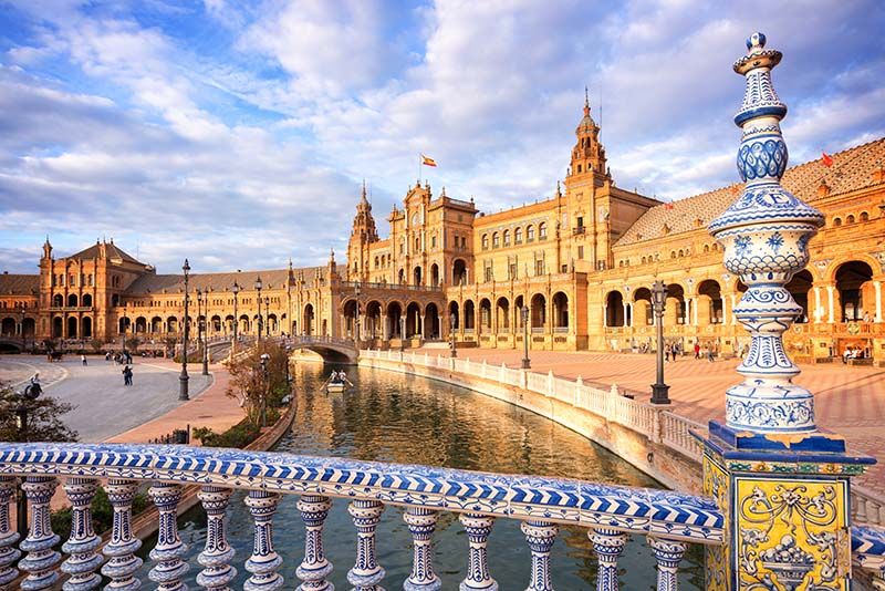 Plaza de España en Sevilla, España, con puentes ornamentados, un canal y un gran edificio bajo un cielo parcialmente nublado.